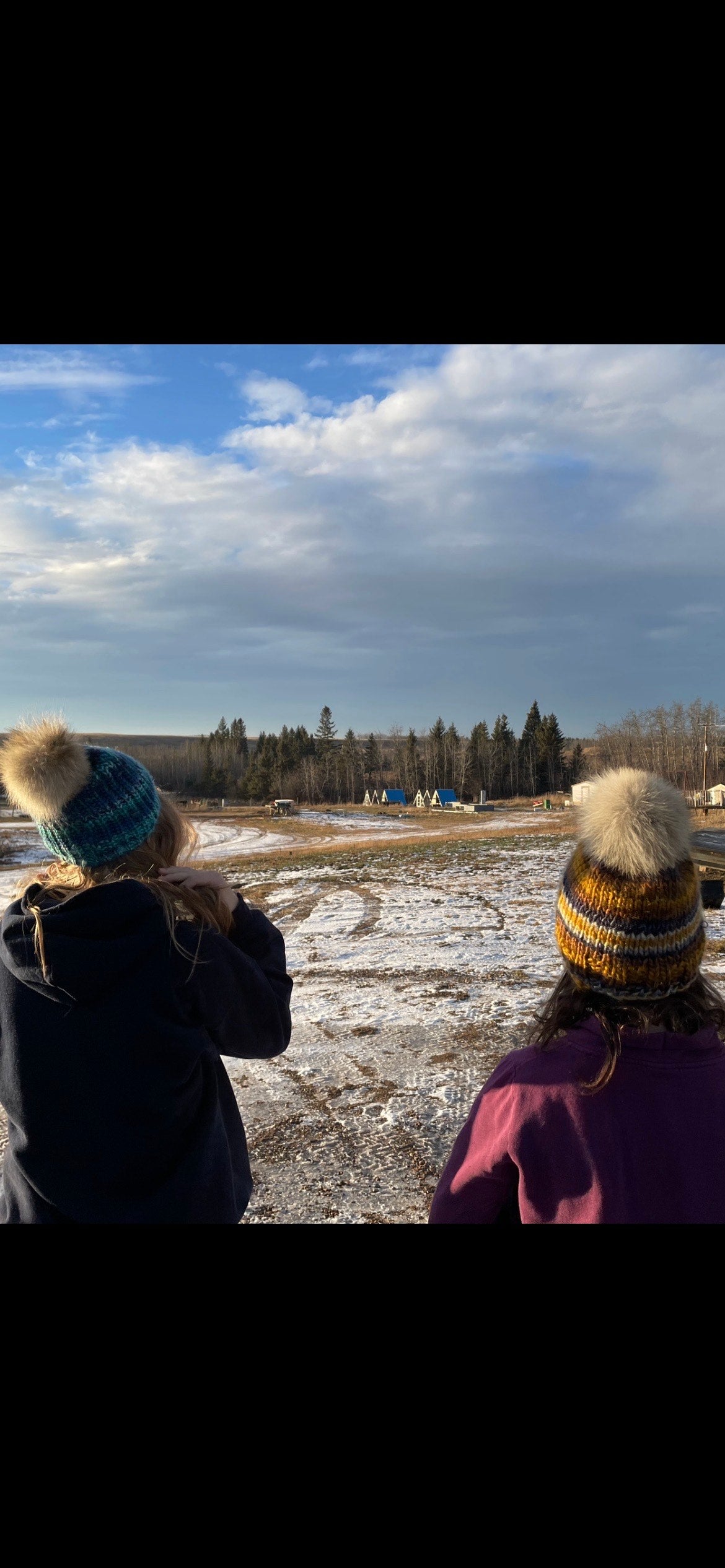 Two people wearing winter hats and scarves looking out over a snowy landscape with trees in the distance.